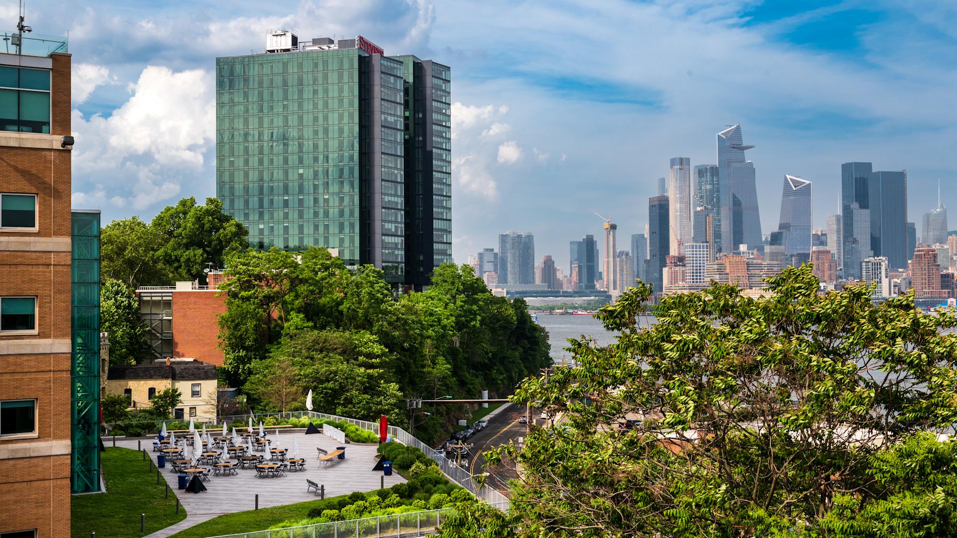 Stevens campus terrace with café tables and umbrellas in summer, the Midtown Manhattan skyline beyond the river