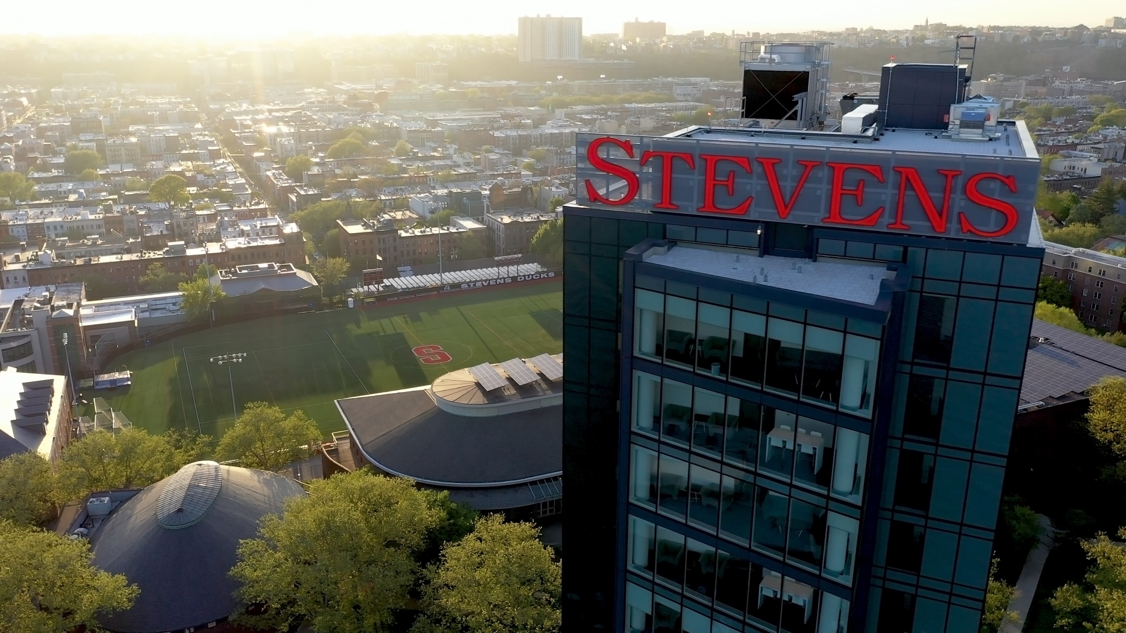 Sunrise aerial of the Stevens tower rooftop sign above the Hoboken campus and athletic field