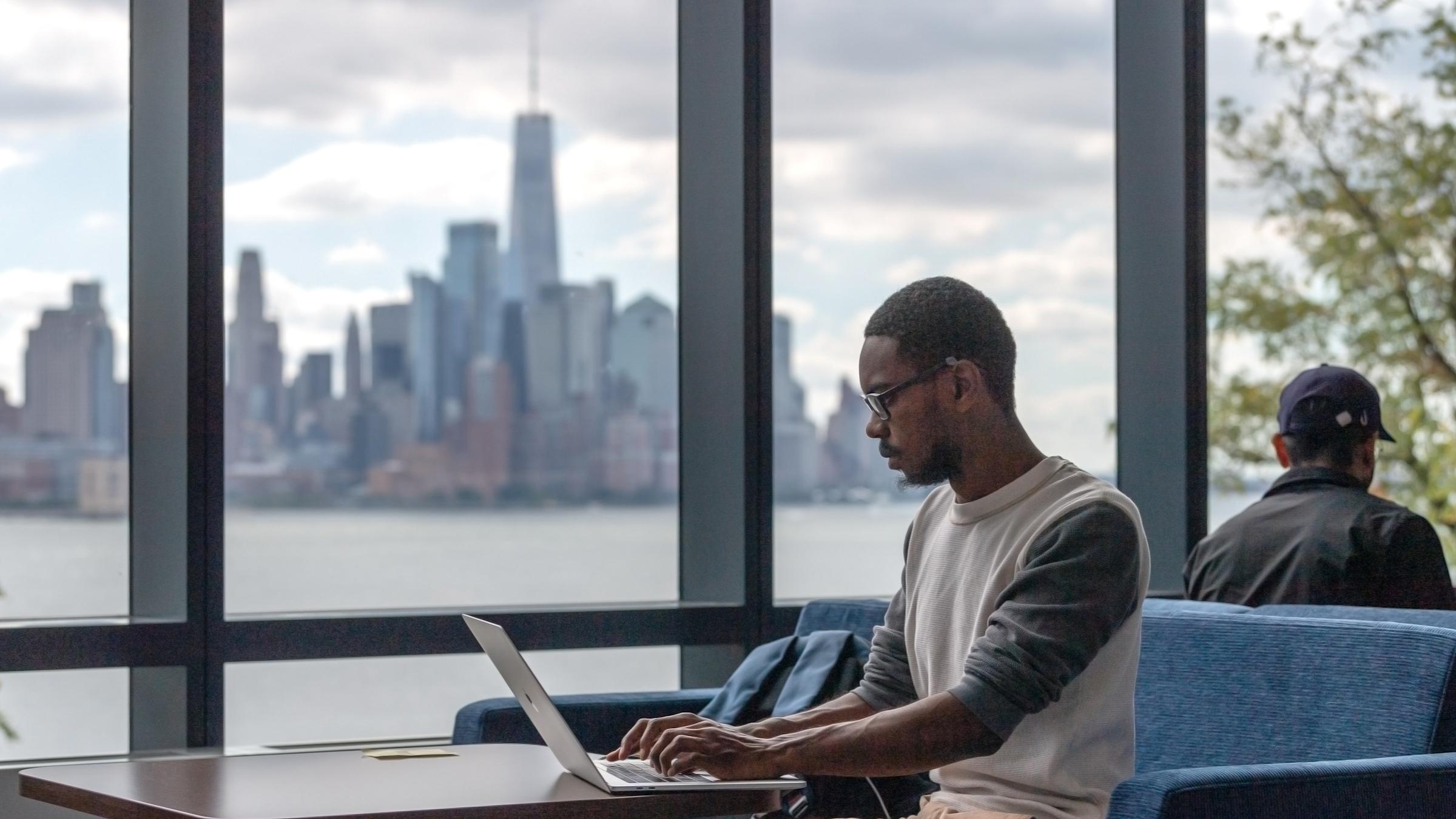 A person working at a table beside floor-to-ceiling windows overlooking the Hudson River and lower Manhattan